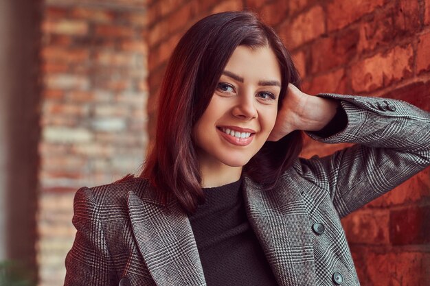 Retrato de primer plano de una chica morena sensual vestida con una elegante chaqueta gris apoyada en una pared de ladrillos en una habitación con interior de loft, mirando a la cámara.