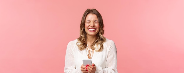 Retrato de primer plano de una chica rubia encantadora, tierna y despreocupada, vestida de blanco, que se ríe de una broma divertida o de un mensaje que sostiene el teléfono móvil con los ojos cerrados y se ríe de algo hilarante de fondo rosa