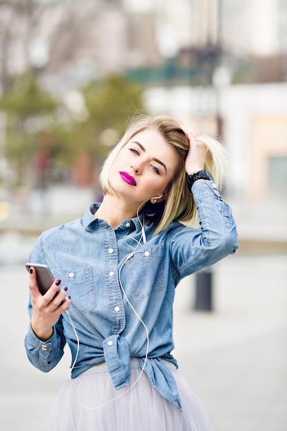 Un retrato de primer plano de una chica rubia de ensueño con labios rosados brillantes escuchando música en un teléfono inteligente