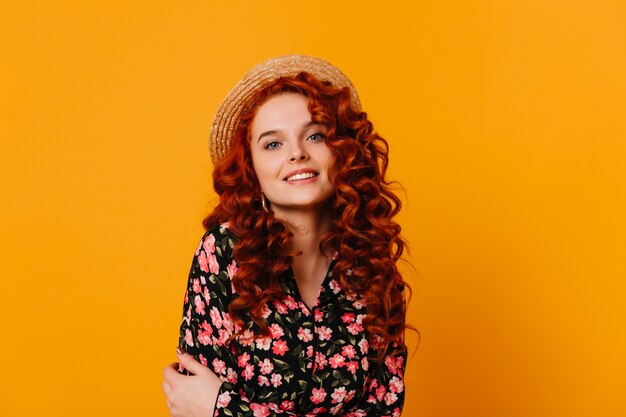 Retrato de primer plano de dama de jengibre con ojos azules brillantes. Chica con sombrero de paja y top estampado floral en espacio naranja.