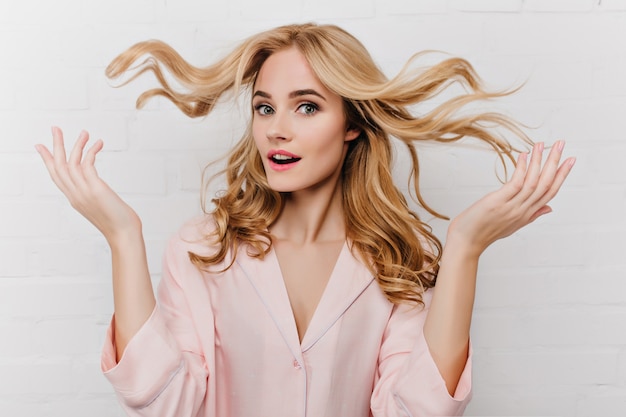 Retrato de primer plano de dama maravillosa juega con su cabello largo rubio. Foto interior de impresionante modelo femenino europeo en pijama rosa aislado en la pared blanca.