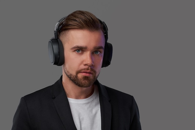 Retrato de primer plano de un elegante hombre barbudo con peinado escuchando música en los auriculares. Aislado en un fondo gris.