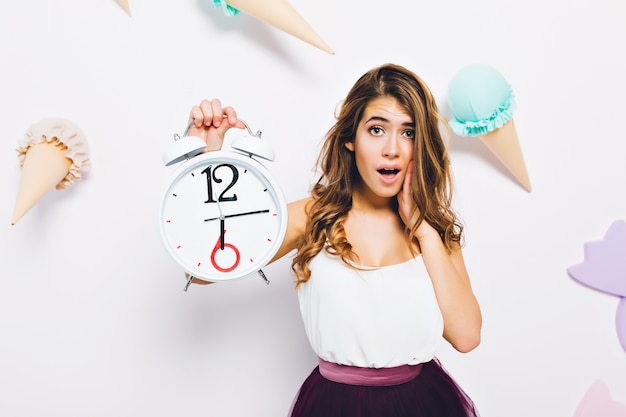 Retrato de primer plano de elegante joven vestida con estilo, posando con reloj blanco en la pared decorada. Chica rizada de pelo largo con expresión de cara infeliz de pie delante de la pared con helado