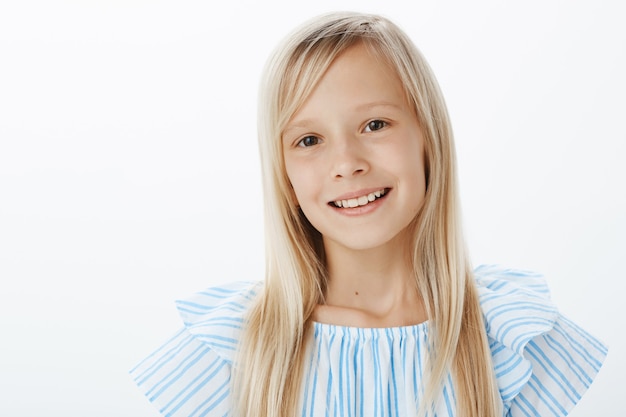 Foto gratuita retrato de primer plano de encantadora adorable niña rubia en blusa azul, sonriendo alegremente y mirando, siendo educado y amigable mientras presenta al nuevo grupo, de pie