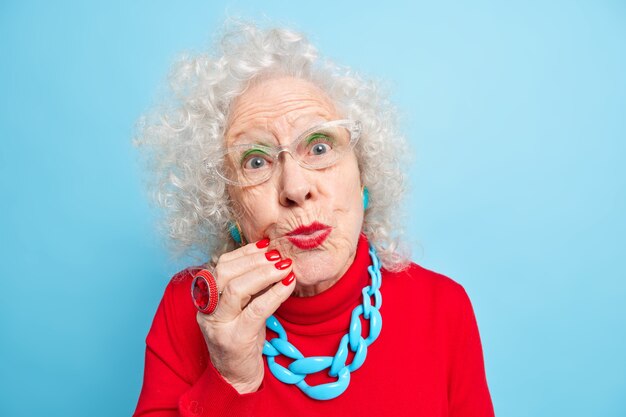 Retrato de primer plano de una encantadora mujer de cabello gris arrugado que mantiene los labios pintados de rojo, se ve doblada con expresión romántica directamente, usa lentes ópticos y un suéter casual con collar