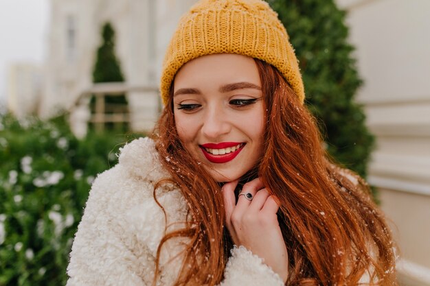 Retrato de primer plano de la encantadora mujer de jengibre de pie cerca de abeto. Foto al aire libre de la alegre niña sonriente con sombrero de invierno.