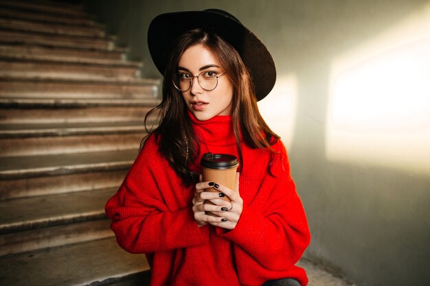 Retrato de primer plano de estudiante morena en suéter rojo y sombrero disfrutando de café en la pared de pasos.