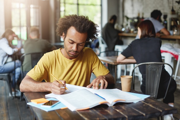 Retrato de primer plano de un estudiante serio de piel oscura con camiseta amarilla sentado en el café durante el descanso tomando café y preparándose para las lecciones de escritura en el cuaderno del libro con lápiz