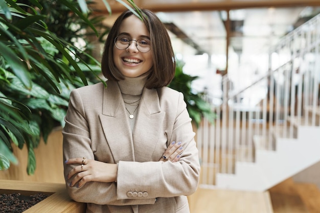 Retrato de primer plano de una exitosa joven sonriente feliz con chaqueta beige y anteojos de pie en el vestíbulo o en la recepción de la oficina saludando al cliente de negocios con una sonrisa agradable invitando a la compañía