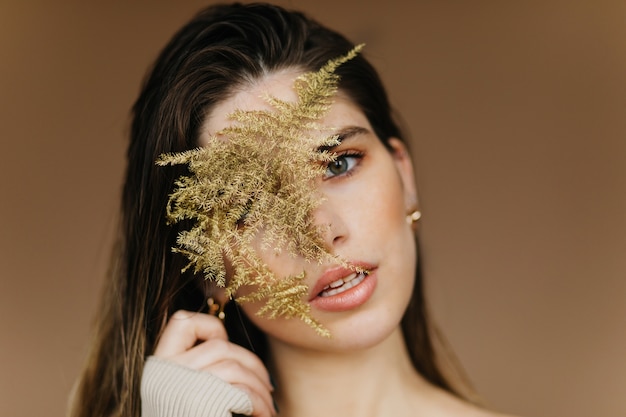 Retrato de primer plano de fascinante chica blanca con planta. Mujer tranquila inspirada de pie en la pared marrón.