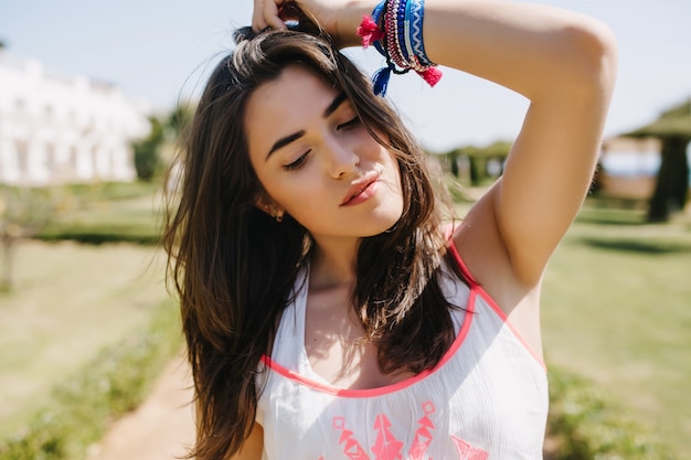 Retrato de primer plano de una fascinante chica morena con accesorios hechos a mano de pie afuera con los ojos cerrados. Encantadora mujer joven con rostro encantador y cabello castaño con camisa blanca posando