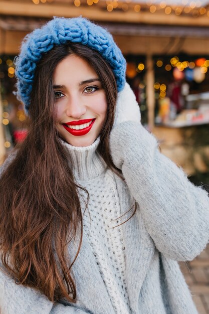 Retrato de primer plano de la fascinante dama de cabello oscuro con ojos verdes esperando la Navidad y riendo. Foto de niña encantadora con peinado largo viste gorro de punto azul y guantes blancos.