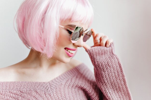 Retrato de primer plano de la fascinante dama de pelo corto con gafas posando en la pared de luz. modelo femenino en peruke rosa de moda.