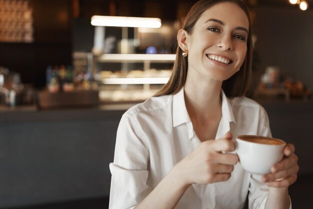 Retrato de primer plano feliz empresaria en blusa blanca, sonriendo alegremente como sentado en un café.