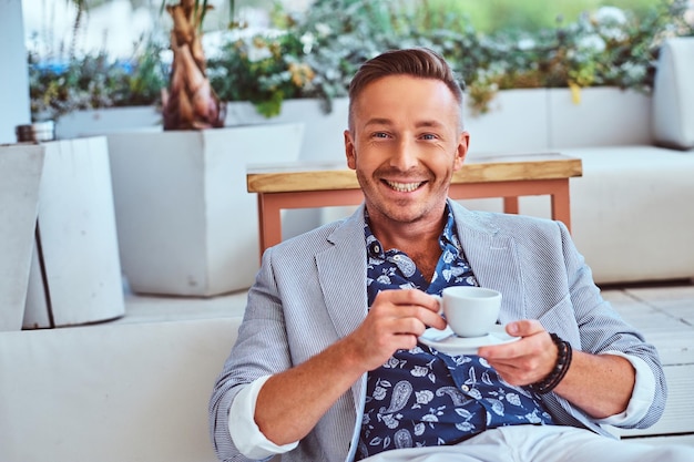 Retrato de primer plano de un feliz hombre rico y elegante vestido con ropa moderna y elegante sostiene una taza de café en un restaurante al aire libre.