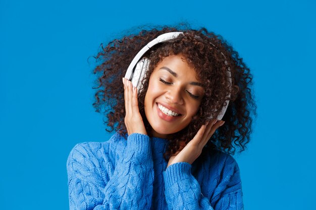 Retrato de primer plano feliz sonriente, romántica y tierna mujer afroamericana disfrutando de escuchar música en auriculares, inclinar la cabeza y cerrar los ojos soñadora y sonriendo encantada, pared azul.