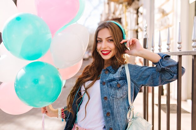 Retrato de primer plano de hermosa chica rizada en chaqueta de mezclilla posando con globos de cumpleaños afuera.