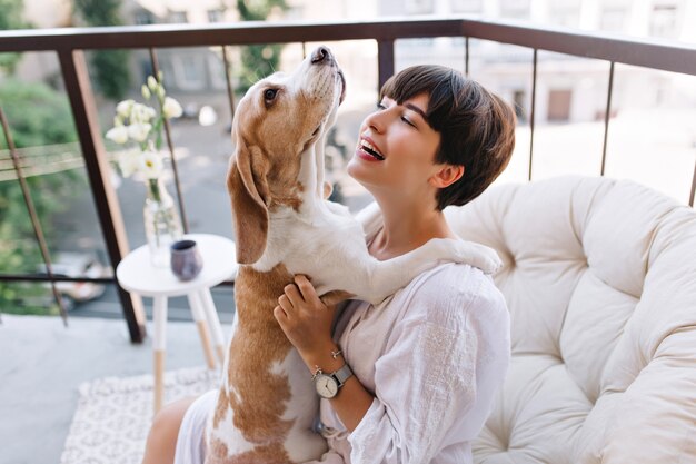 Retrato de primer plano de la hermosa dama de pelo negro mirando con una sonrisa al perrito gracioso mientras está sentado en el balcón. Impresionante chica en bata de baño lleva pulsera y reloj de pulsera jugando con perro beagle