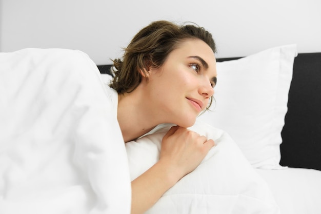 Foto gratuita retrato en primer plano de una hermosa joven acostada en la cama bajo un edredón blanco mirando el reloj despertador