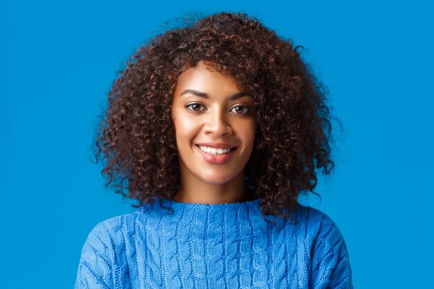 Retrato de primer plano hermosa joven afroamericana con corte de pelo rizado, afro, sonriendo con expresión feliz y agradable, disfrutando de las vacaciones de invierno, vistiendo suéter, pared azul.