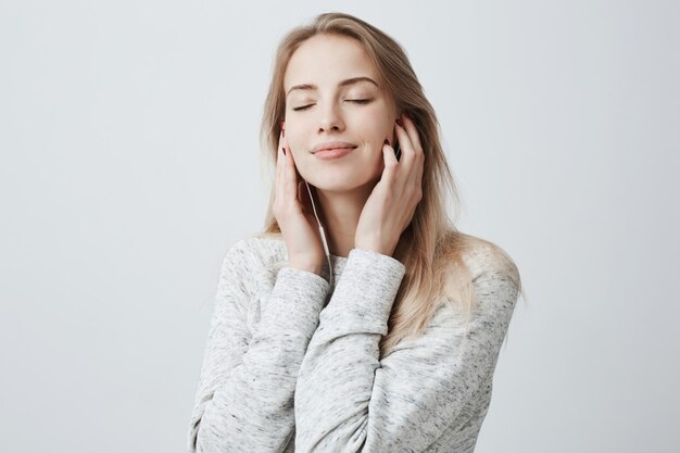 Retrato de primer plano de una hermosa joven europea atractiva en un suéter gris suelto, relajándose con los ojos cerrados, escuchando sus canciones favoritas a través de auriculares blancos, usando la aplicación de música.