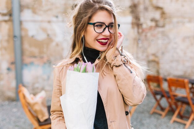 Retrato de primer plano de una hermosa joven en fecha en el café al aire libre con hermosas flores. Encantadora chica con ramo de tulipanes espera amigo en el restaurante mirando a su alrededor a través de gafas