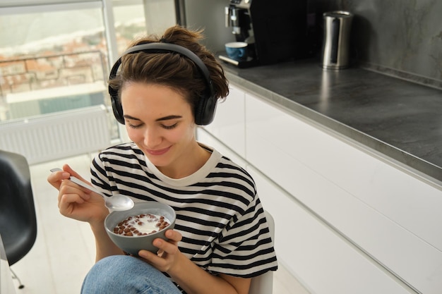 Retrato en primer plano de una hermosa joven sonriente con auriculares comiendo cereales para el desayuno y