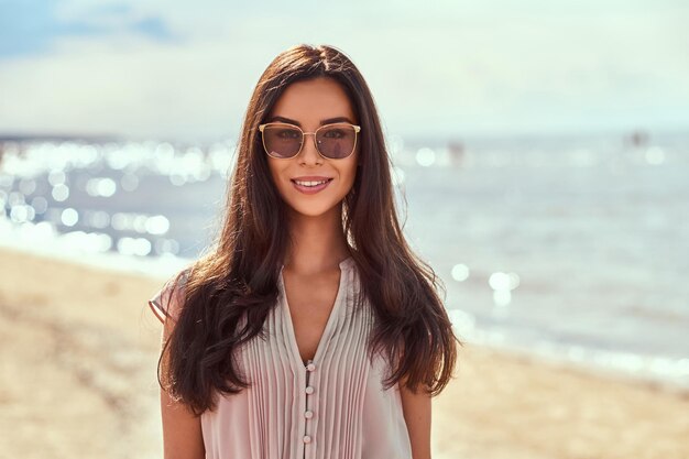 Retrato de primer plano de una hermosa morena feliz con el pelo largo en gafas de sol y vestido en la playa.