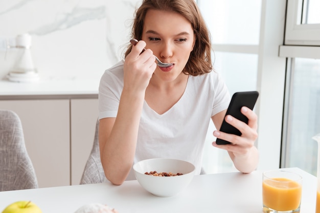 Retrato de primer plano de hermosa mujer hablando por teléfono móvil mientras come copos de maíz en la mesa de la cocina