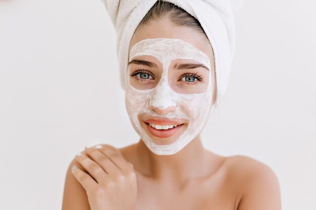 Retrato de primer plano de hermosa mujer joven sonriendo con toallas después de tomar el baño hacer mascarilla cosmética en su rostro.