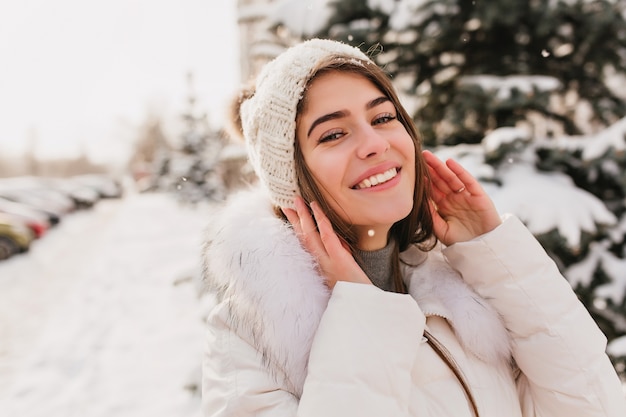 Retrato de primer plano de hermosa mujer con ojos azules posando en la calle en día de invierno cubierto de nieve. Foto al aire libre del encantador modelo femenino en gorro de punto riendo