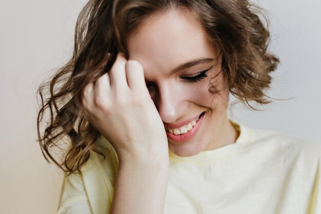 Retrato de primer plano de hermosa niña caucásica posando con sonrisa tímida. Foto interior de una dama de pelo corto tocándose la cara y riendo con los ojos cerrados.