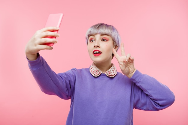 Retrato de primer plano de hermosa niña dollish con cabello corto violeta claro con suéter lila haciendo selfie sobre pared rosa