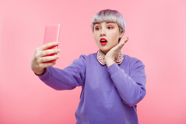 Retrato de primer plano de hermosa niña dollish con cabello corto violeta claro con suéter lila haciendo selfie sobre pared rosa