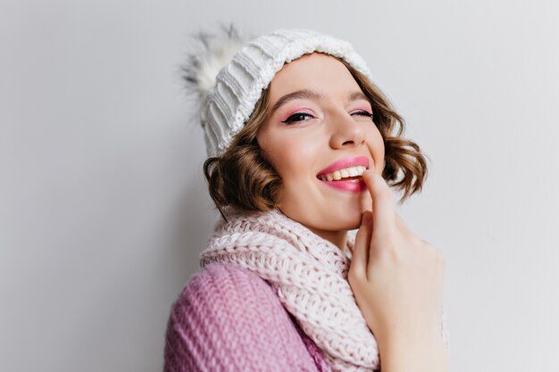 Retrato de primer plano de hermosa niña posando juguetonamente con sombrero de invierno. Foto interior de mujer morena de ensueño en bufanda de pie en la pared de luz.