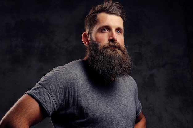 Foto gratuita retrato de primer plano de un hipster con barba completa y corte de pelo elegante, vestido con una camiseta gris, se encuentra en un estudio sobre un fondo oscuro.