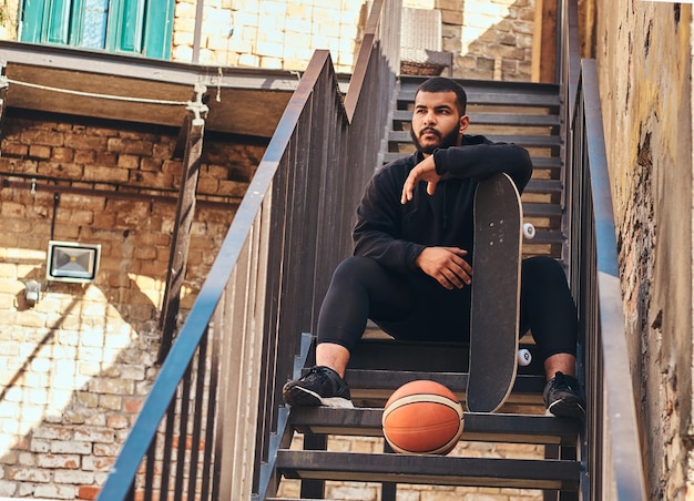 Retrato de primer plano de un hipster barbudo afroamericano vestido con una capucha negra y pantalones cortos deportivos sostiene una patineta mientras se sienta en las escaleras en un gueto.