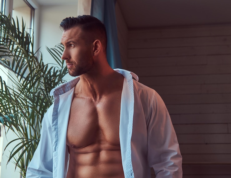 Foto gratuito retrato de primer plano de un hombre alto y barbudo con un cuerpo musculoso, vestido con una camisa blanca y jeans, mirando por la ventana.