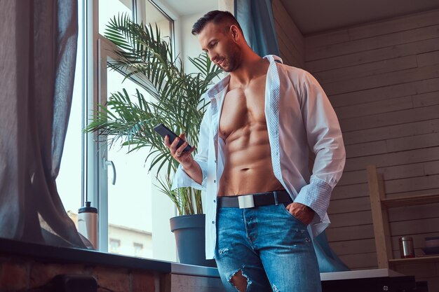 Retrato de primer plano de un hombre alto y barbudo con un cuerpo musculoso, vestido con una camisa blanca y jeans, usando un teléfono inteligente.