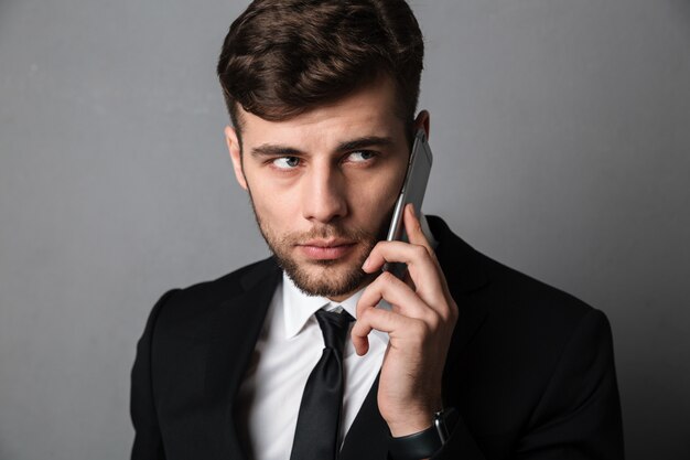 Retrato de primer plano de hombre atractivo joven serio en traje negro hablando por teléfono móvil, mirando a un lado