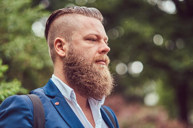 Retrato de primer plano de un hombre barbudo con un corte de pelo vestido con una camisa y una chaqueta con una mochila, de pie en un parque.