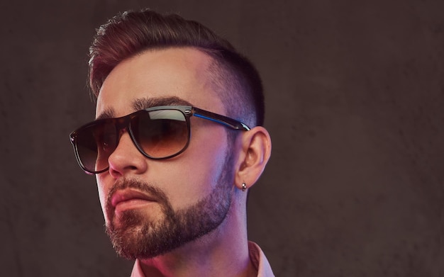 Retrato de primer plano de un hombre barbudo elegante y confiado con peinado y gafas de sol en un traje gris y camisa rosa, posando en un estudio. Aislado en un fondo gris.