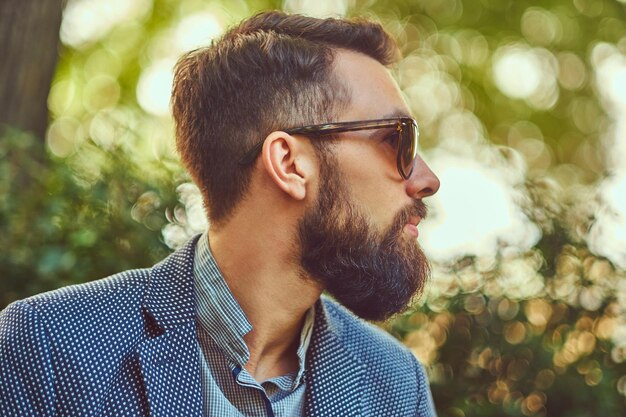 Retrato de primer plano de un hombre barbudo con un elegante corte de pelo, con una chaqueta y gafas de sol, sentado en un banco en un parque de la ciudad.