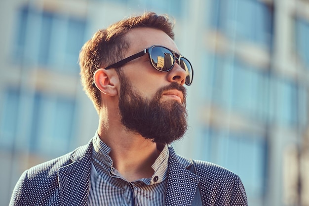 Retrato de primer plano de un hombre barbudo con ropa informal y gafas de sol, parado en una calle de la ciudad contra un rascacielos.