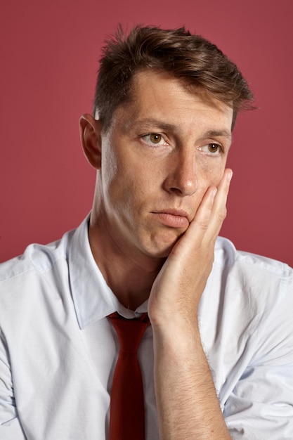 Foto gratuita retrato de primer plano de un hombre brunet atlético con ojos marrones, vestido con una camisa blanca y una corbata de terracota. mira hacia otro lado mientras posa en un estudio con un fondo rojo. concepto de gesticulación