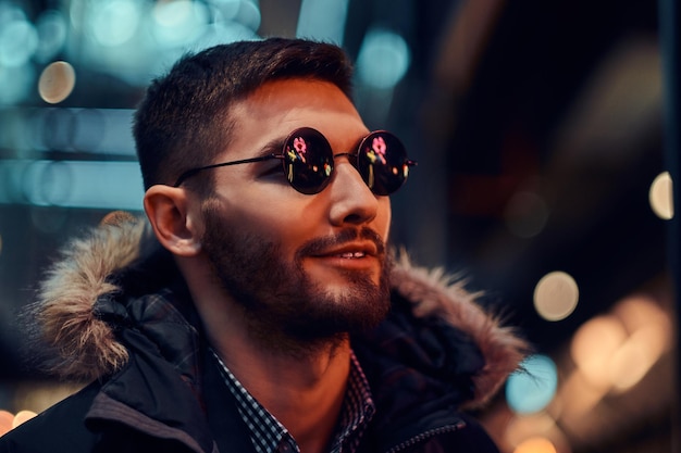Retrato de primer plano de un hombre guapo con un abrigo y gafas de sol de pie en la noche en la calle.