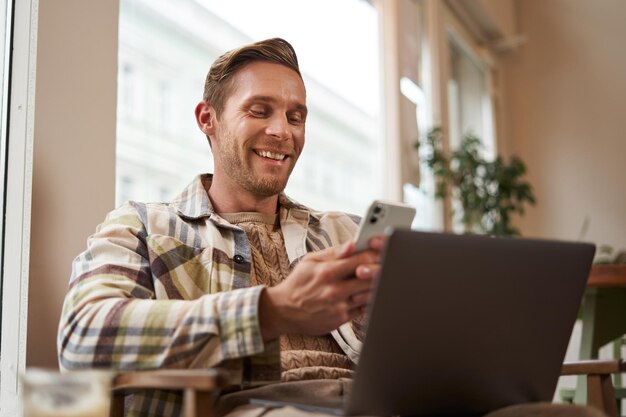 Retrato en primer plano de un hombre guapo y sonriente con un visitante de un café portátil sentado en una silla usando un teléfono inteligente