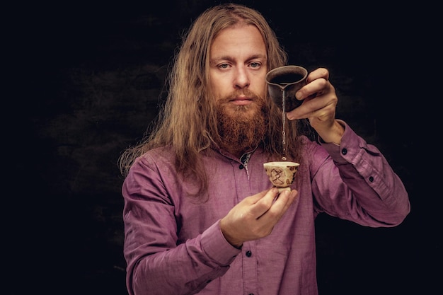 Retrato de primer plano de un hombre hipster pelirrojo con cabello largo y barba llena vestido con una camisa morada preparar té, usando una pequeña tetera y una taza hechas a mano. aislado en un fondo de textura oscura.