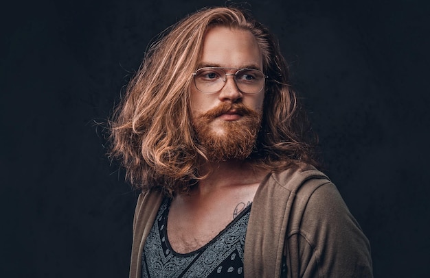 Retrato de primer plano de un hombre hipster pelirrojo con cabello largo y exuberante y barba completa vestido con ropa informal de pie en un estudio, mirando hacia otro lado. Aislado en un fondo oscuro.