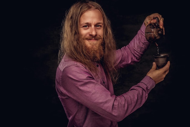 Retrato de primer plano de un hombre hipster pelirrojo sonriente con cabello largo y barba llena vestido con una camisa púrpura preparar té, usando una pequeña tetera y una taza hechas a mano. Aislado en un fondo de textura oscura.